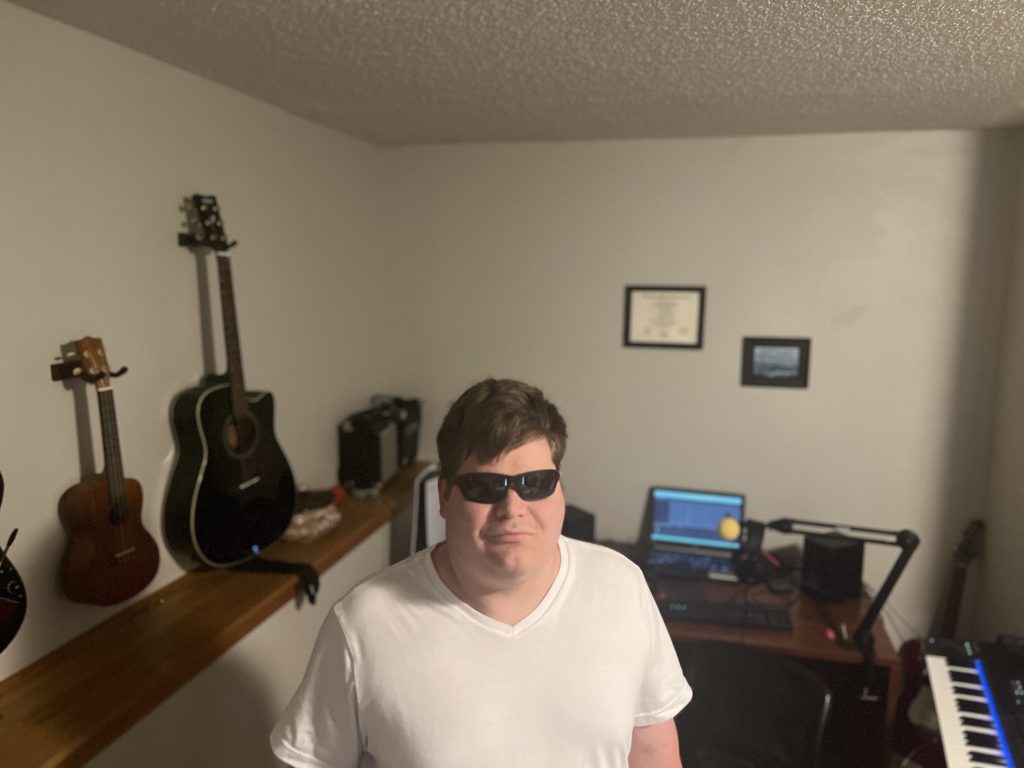 Michael standing in his studio wearing a plain white t-shirt. Behind him there are multiple guitars and a ukulele hanging on the wall. On the right of the photo, there is a midi keyboard as well as a computer displaying a music editing software.